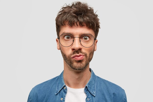 Headshot of puzzled bearded young man looks in wonderment, curves lips, has widely opened eyes, dark wavy hair, recieves unexpected news, thinks over something