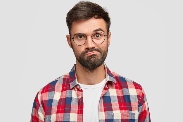 Headshot of puzzled hesitant displeased unshaven young guy purses lips, has uncertain exression, trendy haircut, wears checkered shirt, isolated over white wall. Facial expressions concept