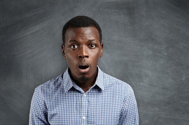 Headshot of puzzled or suprised African employee dressed in checkered shirt looking in shock and frustration against blank chalkboard with copy space for your text or advertising content