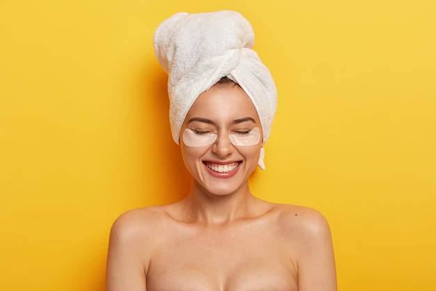 Headshot of satisfied beautiful naked woman applies white patches under eyes to reduce dryness, has pampering session, plumps up skin, wears white towel on head after shower