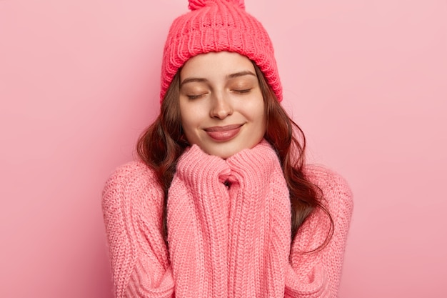Headshot of satisfied European woman has healthy skin, keeps eyes shut, hands under chin, wears warm hat and oversized sweater, isolated over pink background.