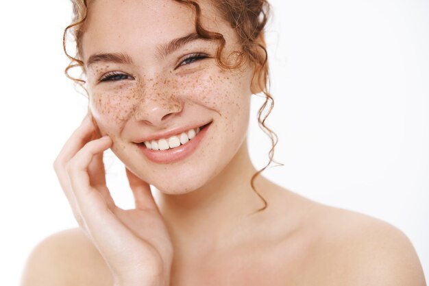 Headshot satisfied tender smiling redhead cute girl freckles curly-haired, standing naked white background laughing happily gently touch cheek look after skin like result apply cosmetology product