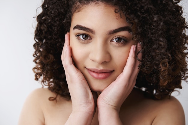 Headshot sensual tender attractive naked european curly-haired brunette female touch pure clean skin cheeks look camera delighted relaxed cute smile take care flaws imperfections, white wall
