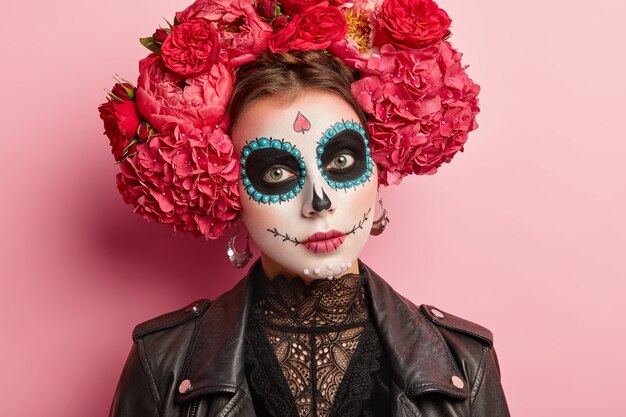 Headshot of serious beautiful woman wears sugar skull makeup, celebrates Mexican Day of dead, wears big earrings, flower wreath, black leather jacket.