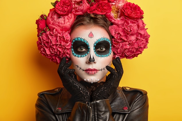 頭蓋骨の化粧をした真面目な美しい女性のヘッドショット、アーティストによって顔を描いた、黒い服を着て、不気味に見えたい、黄色の背景に対してポーズ。伝統的なメキシコの休日