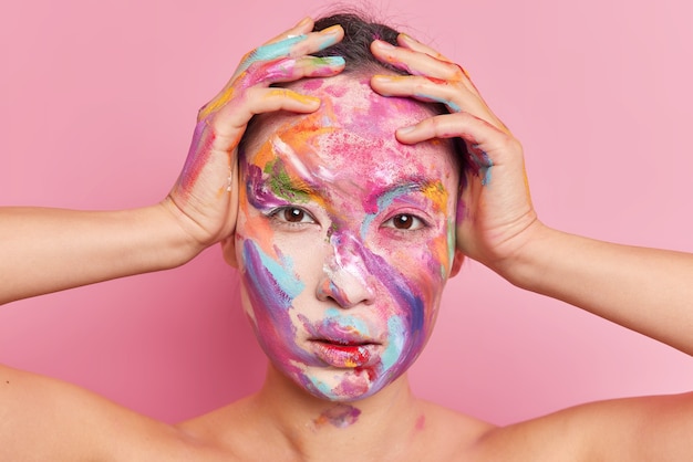 Headshot of serious brunette Asian woman keeps hands on head looks directly at camera smeared with paints poses with bare shoulders against pink background