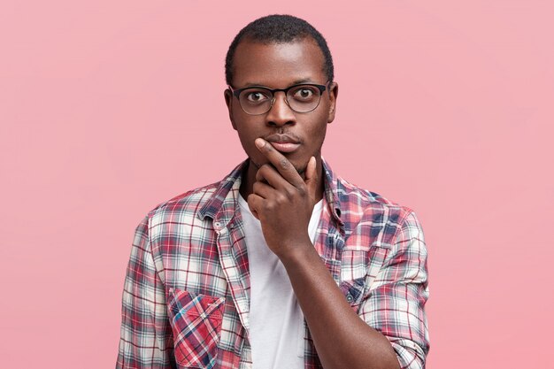 Headshot of serious dark skinned male looks scrupulously at camera, being confident, wears spectacles and checkered shirt, isolated over pink