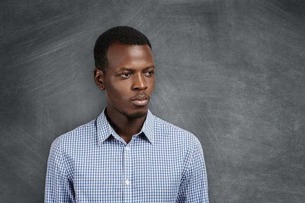 Headshot of serious pensive young African math teacher in casual shirt looking aside with thoughtful expression, standing at blackboard in classroom, waiting for his students for next lesson