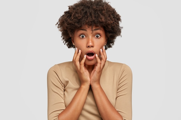 Headshot of shocked beautiful black woman opens mouth widely, keeps both hands on cheeks