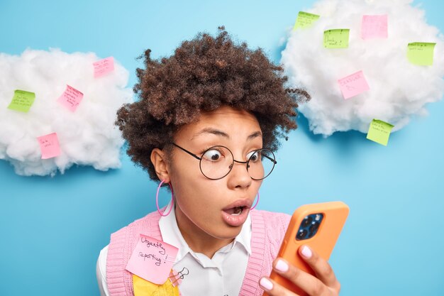 Headshot of shocked curly haired woman stares at smartphone screen finds out she failed exam has much work to do makes reminding notes on stickers prepares report urgently isolated on blue wall