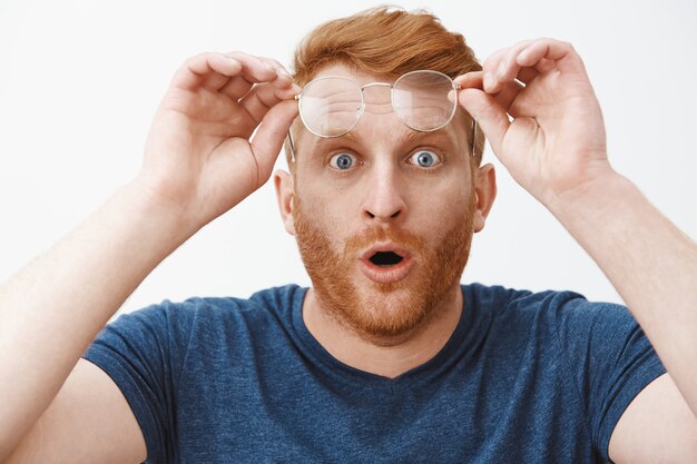 Headshot of shocked and impressed overwhelmed redhead guy with beard, taking off glasses and holding rim on forehead, folding lips and staring with popped eyes at impressive and curious thing