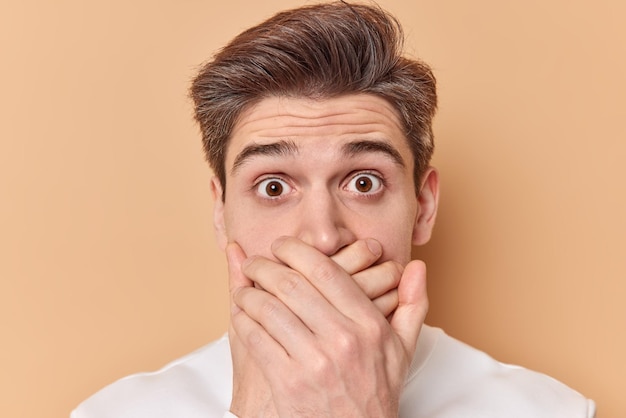 Headshot of shocked impressed young European man covers mouth with hands stands speechless tries to be speechless reacts on amazing news isolated over beige background. Human reactions concept