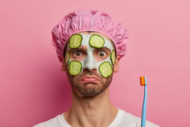 Headshot of shocked man cleanses facial skin, holds toothbrush, shower cap, ready for cleaning teeth, has serious strict look, models against pink space