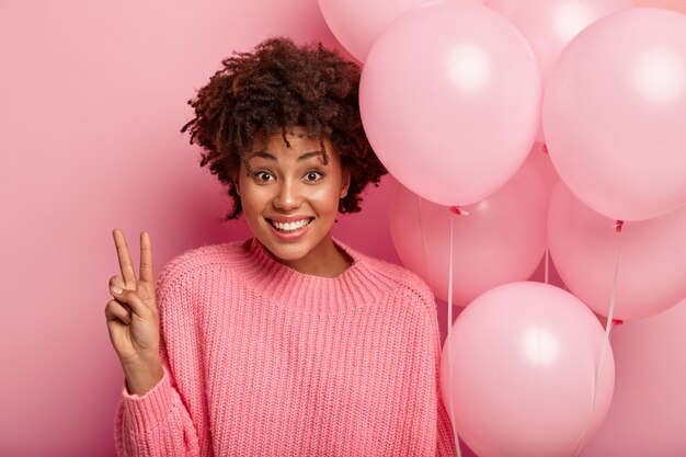 Headshot of smiling female shows peace gesture, wears oversized knitted jumper, holds air balloons, poses for making photo, celebrates festive event, isolated over rosy wall.