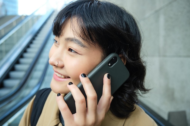 Free photo headshot of smiling korean woman with smartphone makes a phone call goes down escalator in city comm