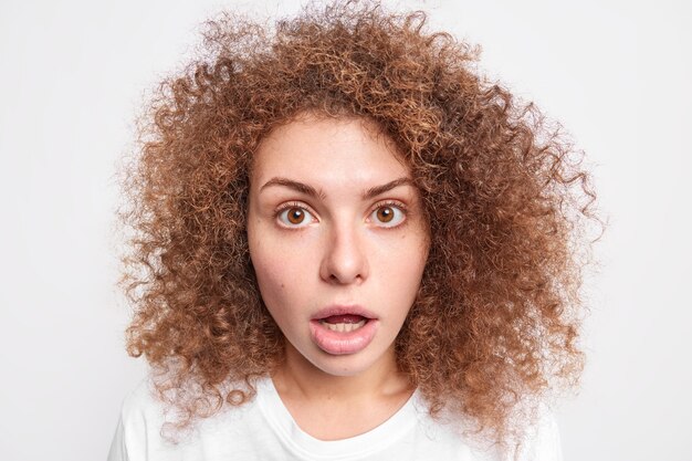 Headshot of startled speechless woman keeps jaw dropped notices something shocking stands in stupor reacts on unexpected revelation has curly hair isolated over white wall. Omg concept