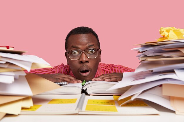 Headshot of stupefied male professor leans head on table, has surprised facial expression, wears eyewear, keeps hands on opened notebook