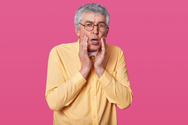 Headshot of stupefied terrified thin man wears yellow shirt, keeps hands on cheeks. Surprised elderly male with glasses against rose wall