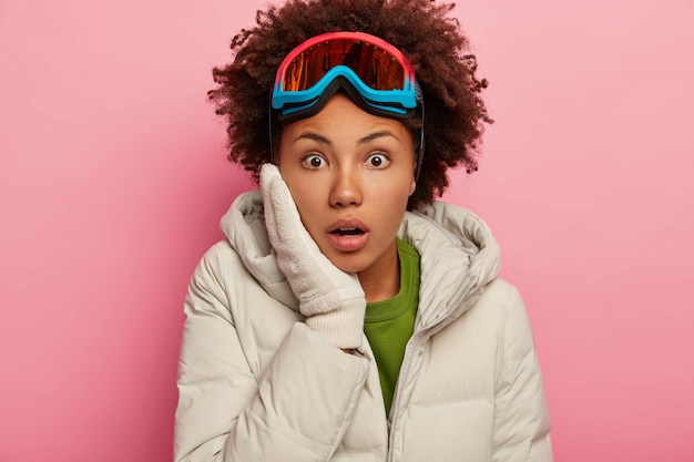 Primo piano del volto di una donna afroamericana sorpresa che tiene la mano sulla guancia, guarda con paura, indossa occhiali da sci e capispalla