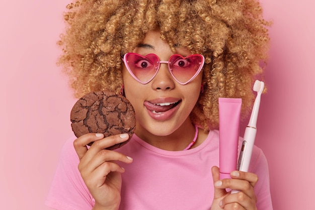 Free photo headshot of surprised curly haired young woman licks lips holds appetizing chocolate cookie toothpaste in tube and toothbrush has wondered expression wears heart shaped sunglasses casual t shirt