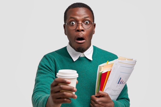 Headshot of surprised dark skinned man holds takeaway coffee, carries papers, surprised to hear shocking news