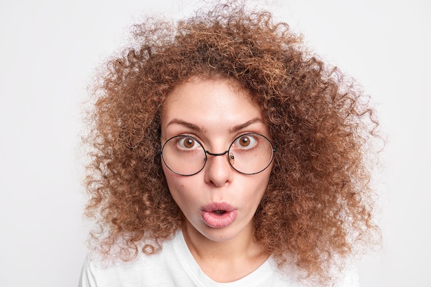 Free photo headshot of surprised european woman with curly bushy hair looks with amazement  cannot believe in shocking news wears round spectacles isolated over white  wall. omg concept