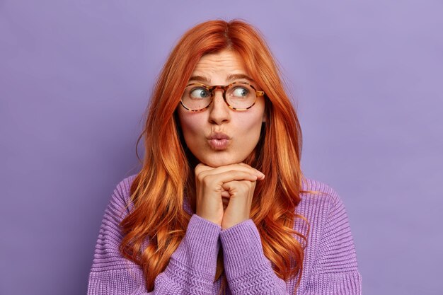 Headshot of surprised ginger young woman keeps lips folded hands under chin looks aside dressed in long sleeved jumper.