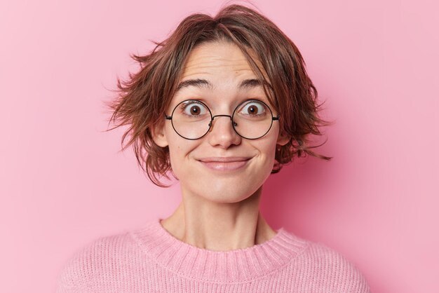 Headshot of surprised happy woman stares through glasses cannot believe in shocking relevation has trendy bob hairstyle wears casual jumper isolated over pink background hears something pleasant