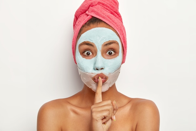 Headshot of surprised young female makes hush hand sign, tells beauty treatment, looks with bugged eyes, applies clay facial mask, silicone patch near lips, has mysterious expression, tells be qiet