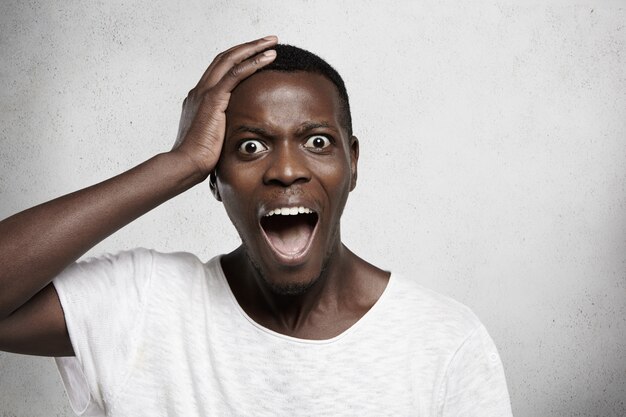 Headshot of terrified young African employee in white t-shirt holding hand on his head and screaming with horrified look, opening mouth widely. Black male feeling scared or stressed.