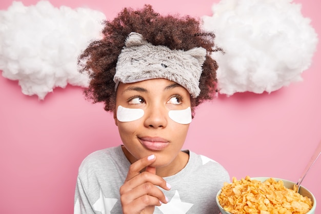 Headshot of thoughtful dreamy curly woman keeps hand on chin looks pensivey aside dressed in pajama has healthy breakfast holds bowl of cornflakes makes plans for working day