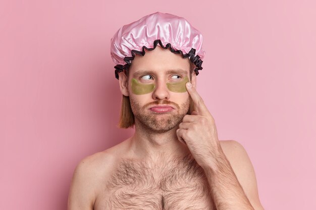 Headshot of unhappy adult European man applies green collagen patches under eyes reduces wrinkles stands shirtless