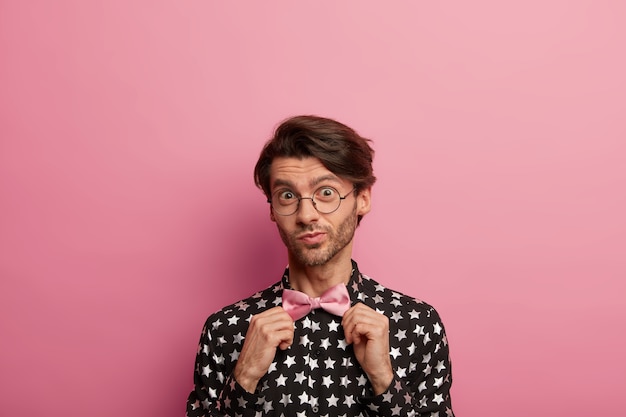 Headshot of unshaven man adjustes rosy bowtie, has elegant look, wears black shirt with star print, looks through round glasses