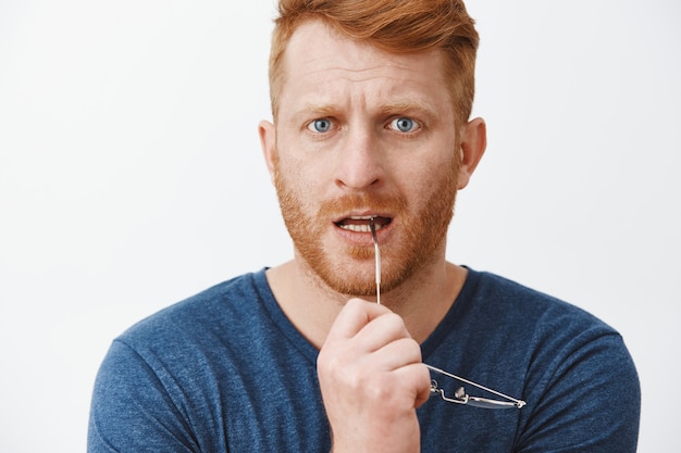 Headshot of worried redhead man with bristle, biting rim of glasses and frowning, looking intense feeling nervous while watching at company chart, standing anxious over gray wall