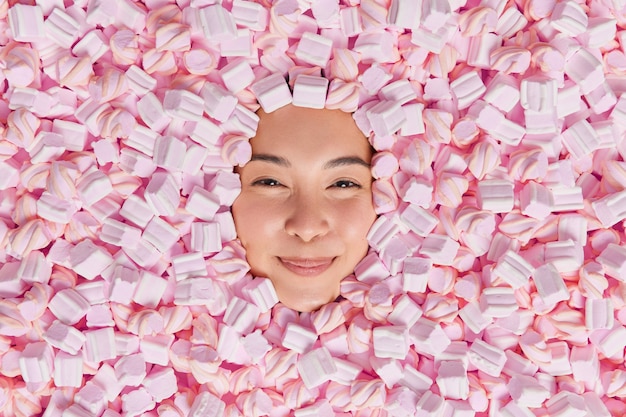 Headshot of young cheerful woman eats marshmallows lies around tasty dessert sticks head consumes excess of calories has sweet tooth enjoys cheat meal day.