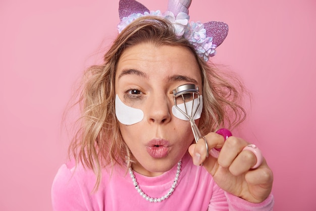 Headshot of young female model uses eyelashes curler while applying makeup keeps lips rounded puts on white patches under eyes for skin moisturising poses against pink wall Beauty time concept