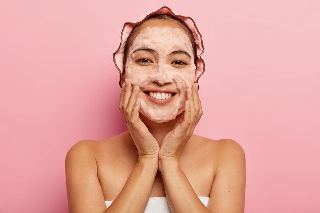 Headshot of young korean female touches flawless soft skin, washes face with hygienic soap with foaming cleanser, wrapped in towel, has bath cap on head, isolated on pink wall. Cleaning concept