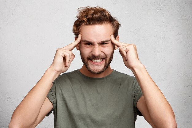 Headshot of young man holds fingers on temples, has bad memory, tries to concentrate and remember