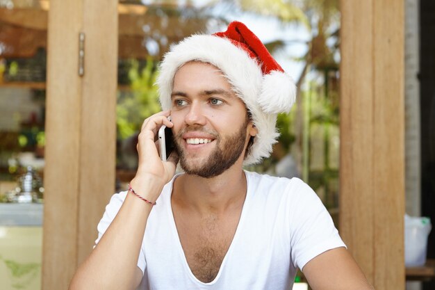 Headshot of young unshaven man dressed in white t-shirt and red Santa Claus hat looking happy while speaking on mobile phone with his girlfriend, listening to her warm congratulations on Christmas