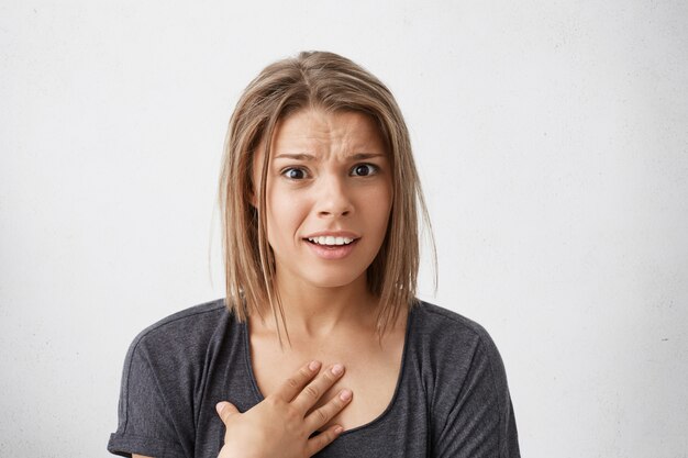 Headshot of young woman holding hand on her breast, verbally defending herself, having perplexed and puzzled expression on her face, saying: Who me? Human emotions, feelings, reaction and attitude