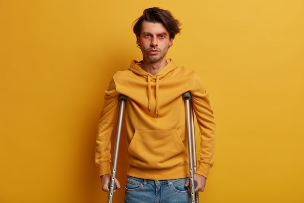 Health care concept. Disabled man with crutches being handicapped after tragic accident, has bruises and abrasion, disable to walk, isolated over yellow wall. Mobility assistance. Injured male