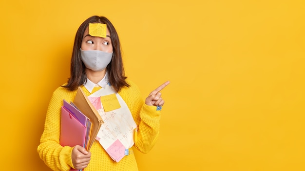 Concetto di assistenza sanitaria e istruzione. la studentessa adolescente felice indossa una maschera protettiva contro i punti del coronavirus su un muro giallo vuoto dimostra i risultati del suo progetto circondati da foglietti adesivi