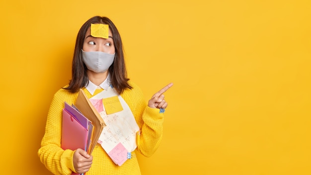 ヘルスケアと教育の概念。満足している10代の学生は、空白の黄色い壁にコロナウイルスのポイントに対する保護マスクを着用し、紙の付箋紙で囲まれた彼女のプロジェクトの結果を示しています
