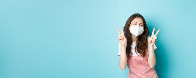 Free photo health coronavirus and social distancing concept happy young woman in medical respirator showing vsigns cute girl posing in face mask with peace gesture blue background