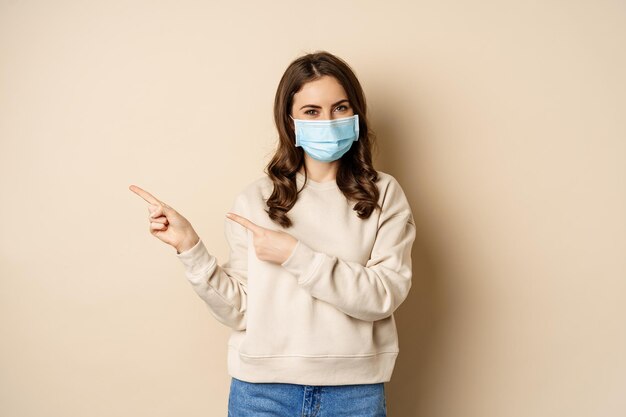 Концепция здоровья и ковид-19. Симпатичная брюнетка в медицинской маске, указывая пальцами на логотип, баннер, показывая пространство для копирования, стоя на бежевом фоне