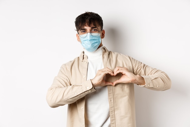 Концепция здравоохранения и карантина, романтический молодой человек в стерильной медицинской маске, показывающий жест сердца ...