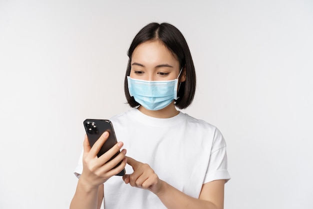 Здоровье и концепция covid19 Молодая азиатка в медицинской маске с помощью мобильного телефона, печатающего на смартфоне, стоящем на белом фоне
