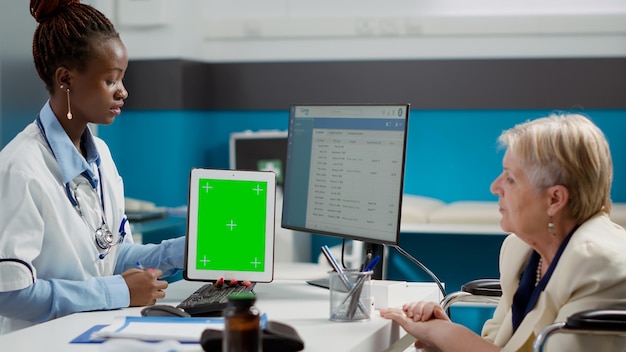 Free photo health specialist showing greenscreen on tablet to woman with disability in cabinet. looking at blank mockup template with isolated display and chroma key copyspace, wheelchair user.