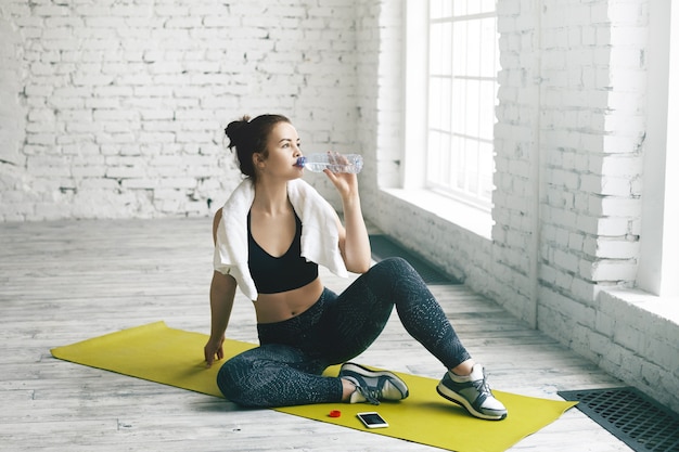 Health, sports, fitness, diet and weight loss concept. Beautiful young brunette female wiping sweat with towel after physical workout, sitting on mat and drinking fresh water out of plastic bottle