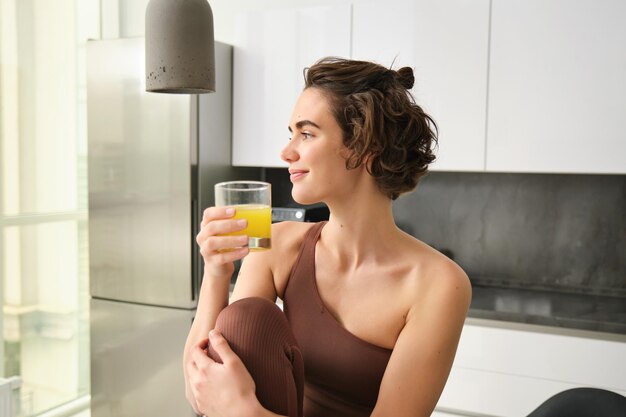 Health and wellbeing young brunette woman drinking glass of orange juice in her kitchen looking at t